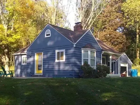 Blue house with white trim and yellow door in a wooded yard, lit by sunlight
