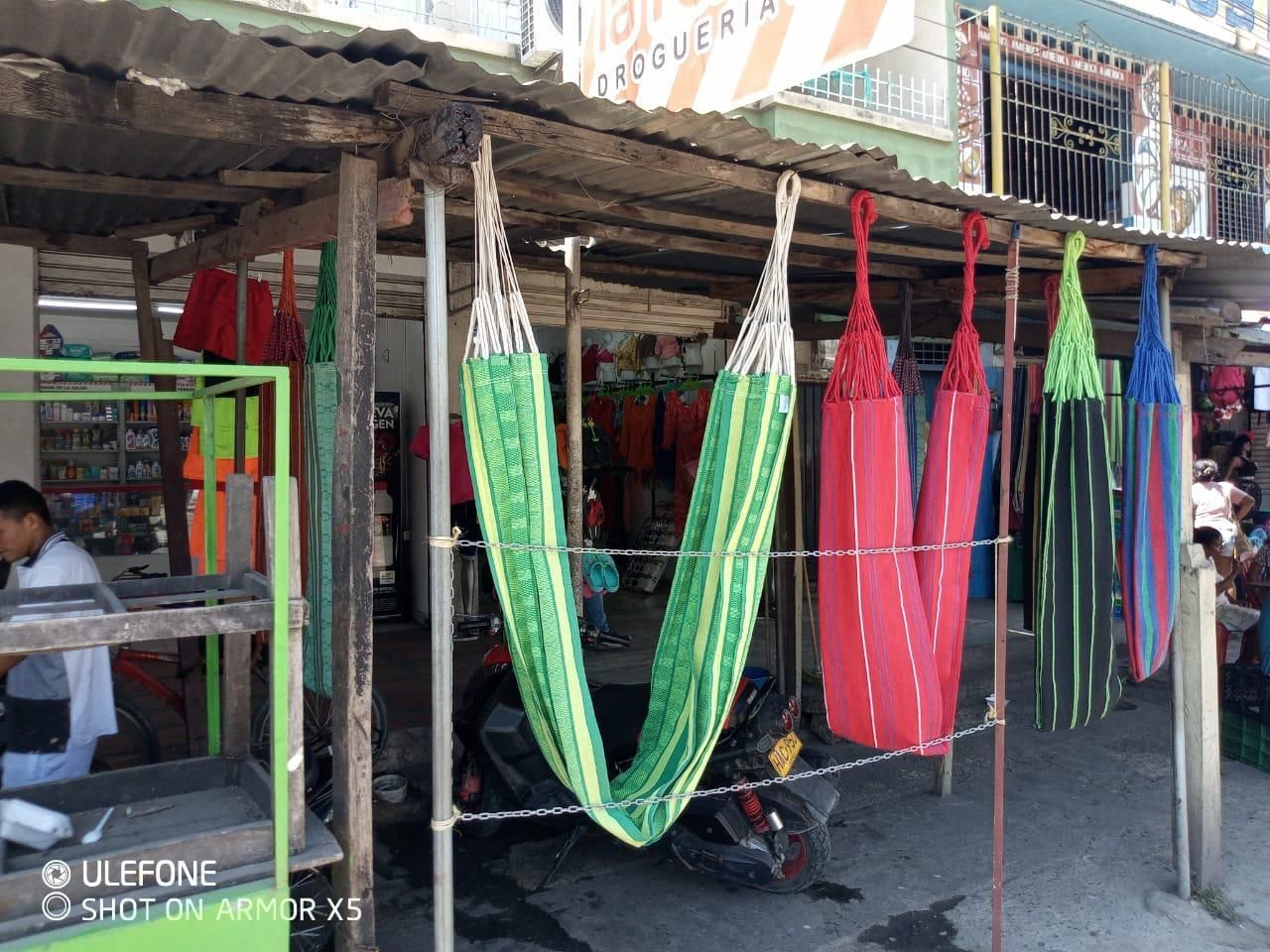 Un puesto de mercado exhibe varias hamacas tejidas de colores que cuelgan de una sencilla estructura de techo de madera.