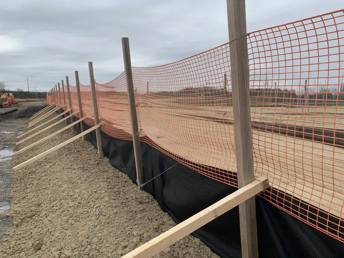 Construction site erosion control fence with orange safety mesh along a gravel slope under cloudy skies