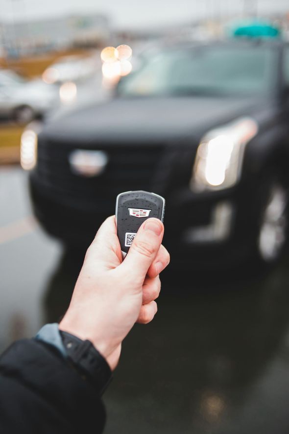 Hand holding car key fob, with black Cadillac SUV in the background, outdoors on a wet day.