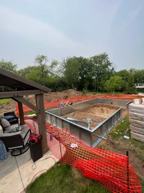 A rectangular swimming pool under construction in a backyard, enclosed by orange safety fencing and near a gazebo.