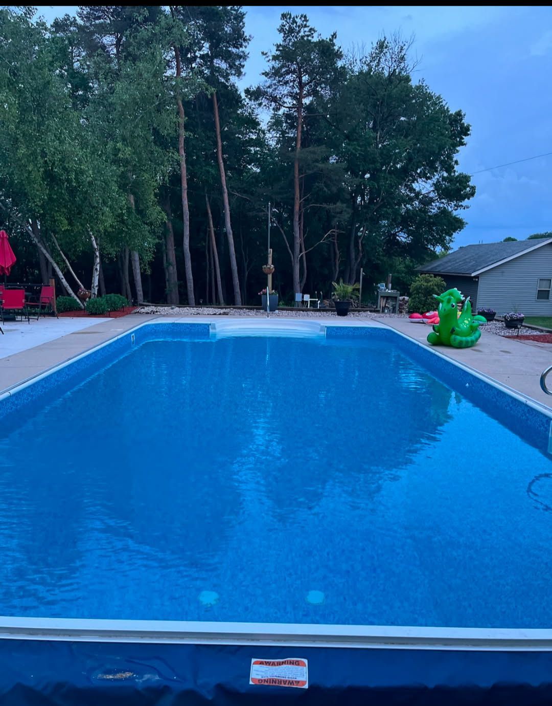A rectangular backyard swimming pool filled with blue water, surrounded by a patio, trees, and a house at dusk.