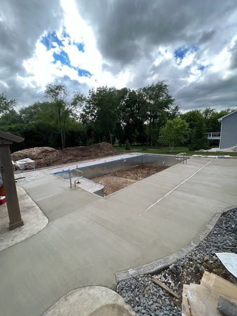 Newly poured concrete pool deck surrounds an empty, partially completed swimming pool under a cloudy sky.