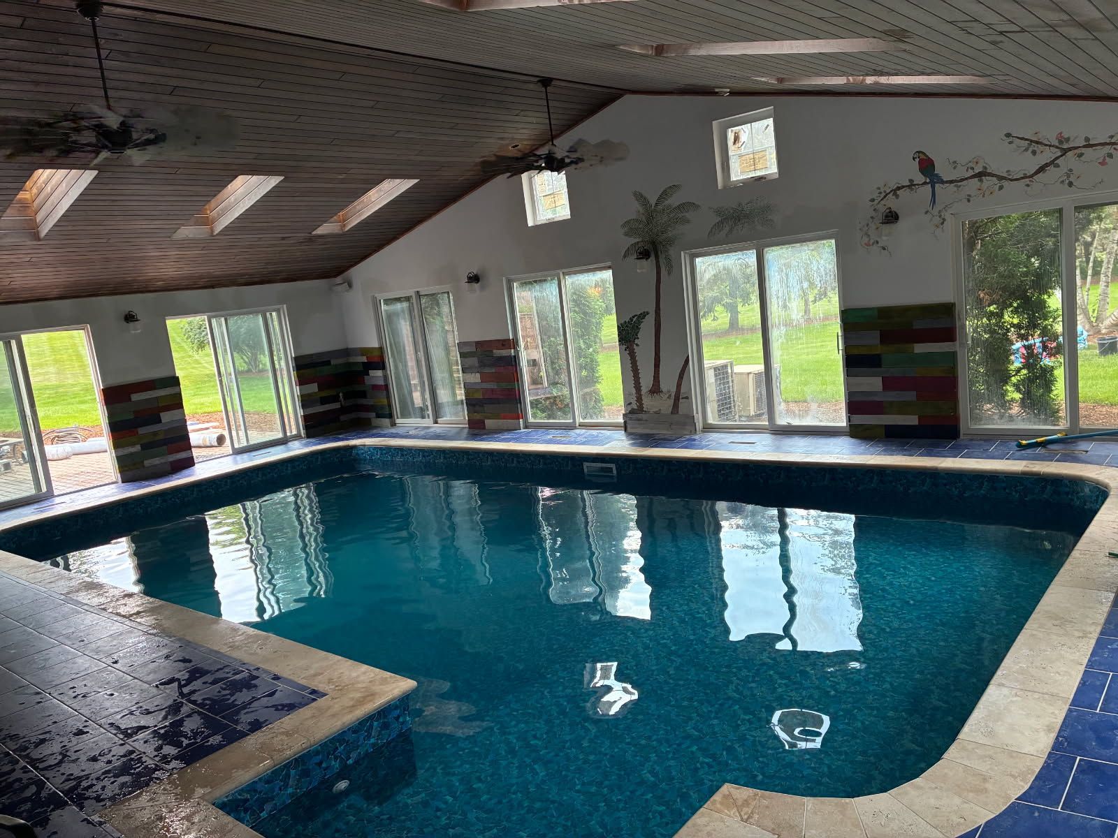 An indoor swimming pool with dark blue tiles, surrounded by light-colored stone and multiple glass sliding doors.