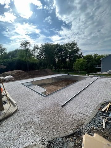 A backyard under construction featuring a rectangular pool frame surrounded by a gravel base on a sunny day.