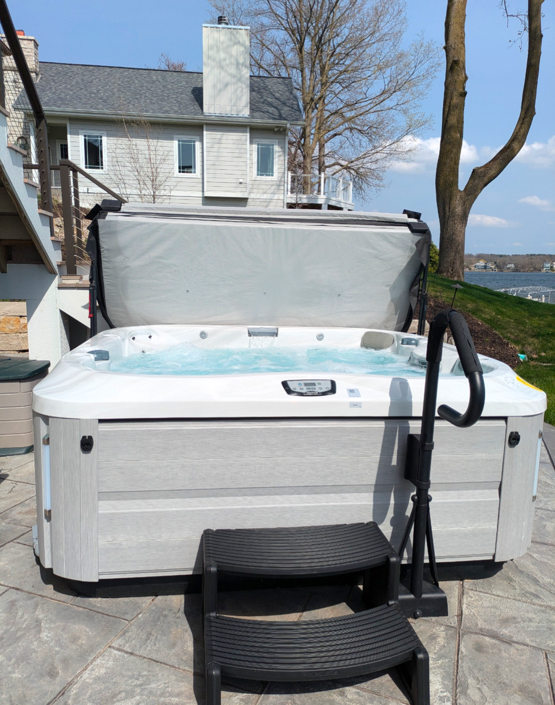 A grey and white hot tub with an open cover sits on a stone patio outside a house near a lake, with black steps and rail.