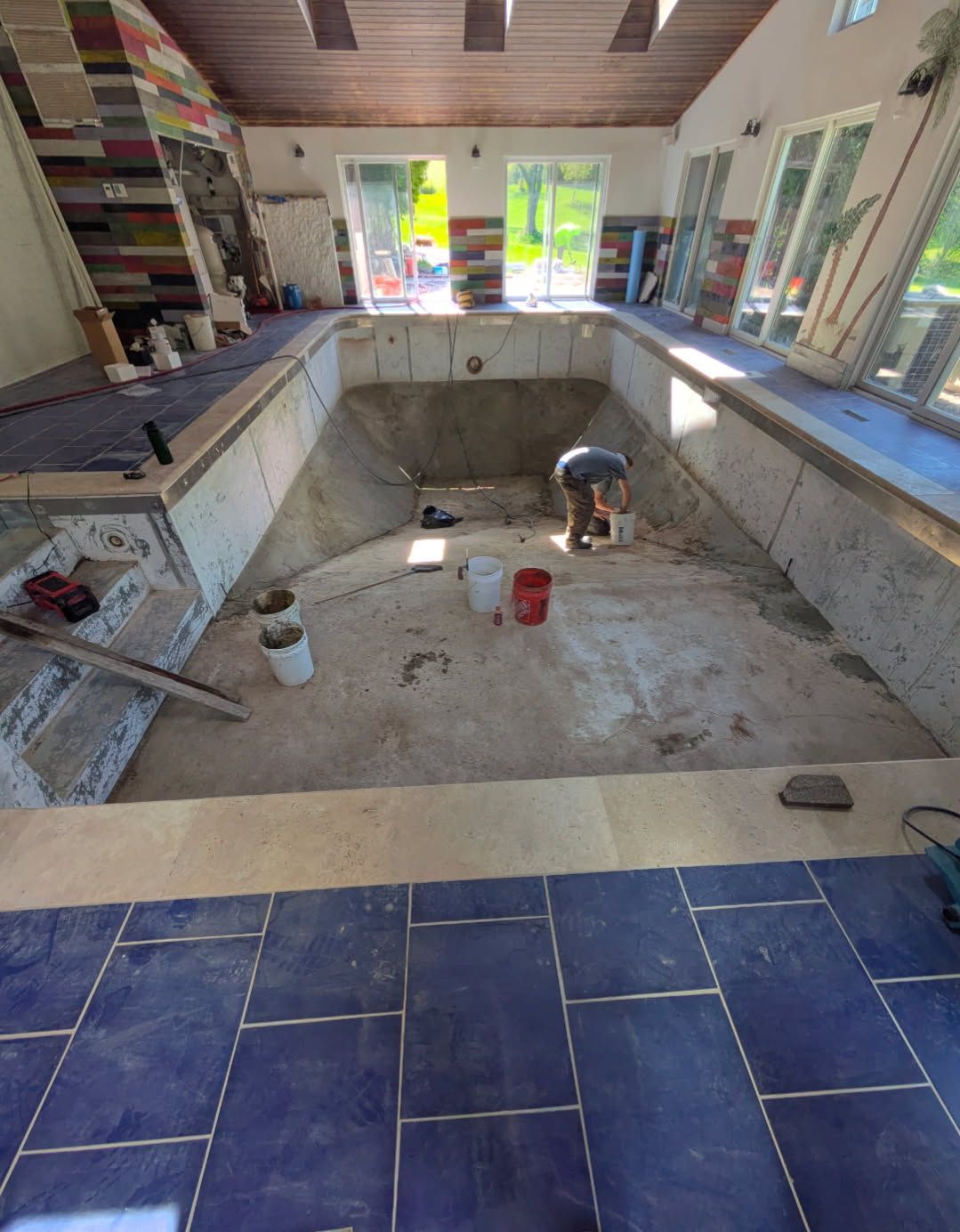 A person working inside an empty, concrete indoor swimming pool under renovation, with buckets nearby.