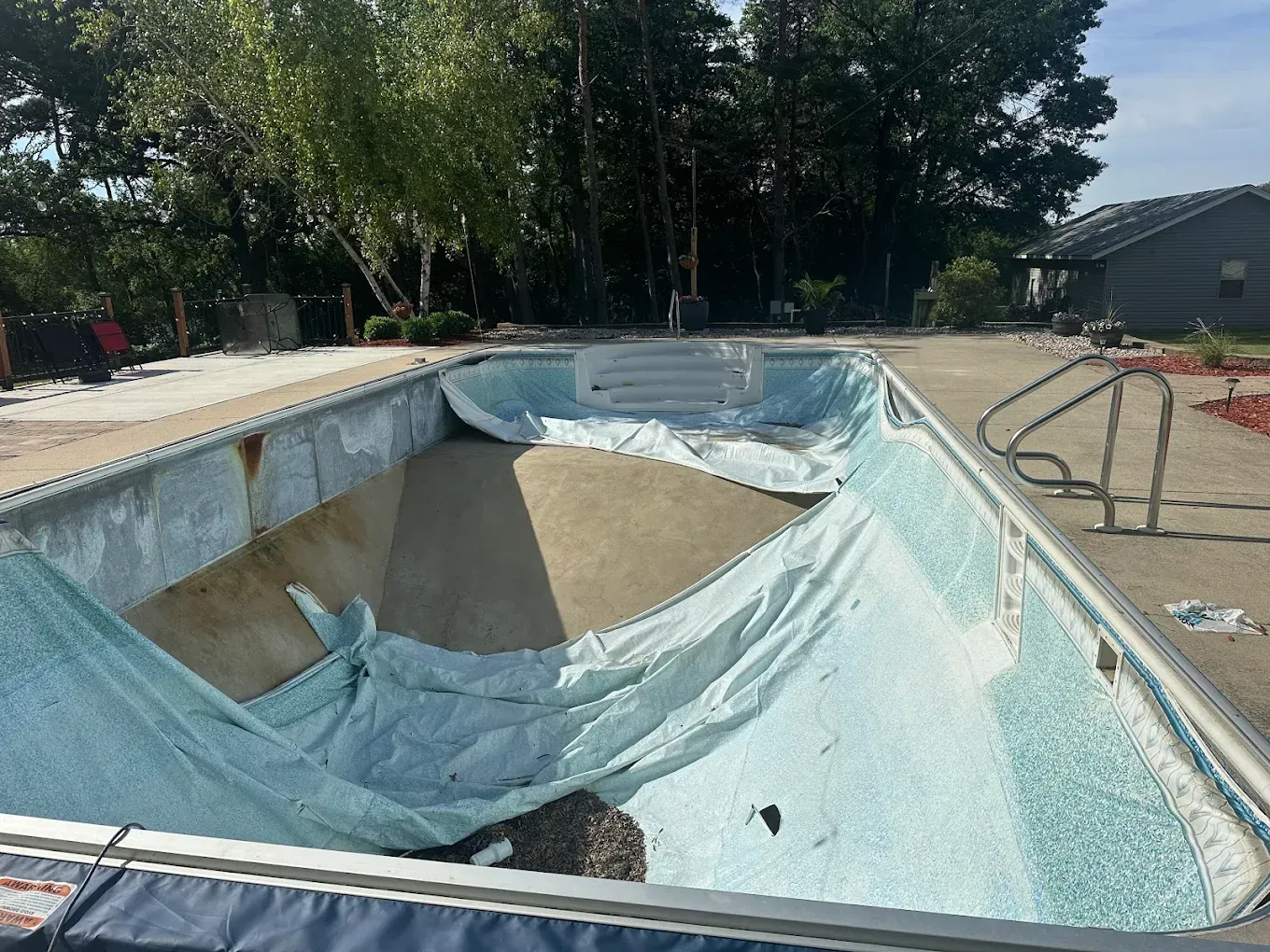 An empty backyard swimming pool with a loose, light blue vinyl liner sagging against the concrete walls and floor.