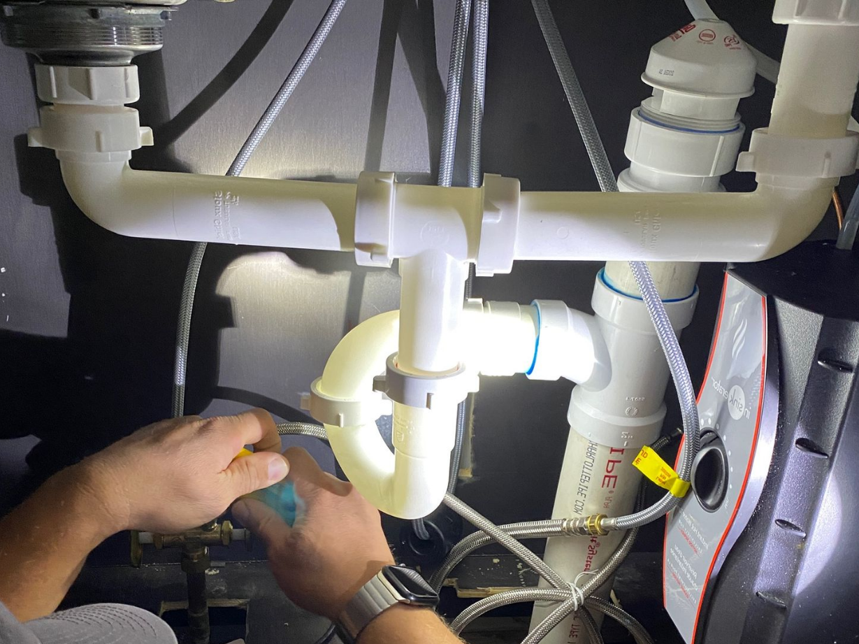 Plumber repairing a sink pipe under a bathroom basin, with tools beside him and two wall-mounted sinks visible.