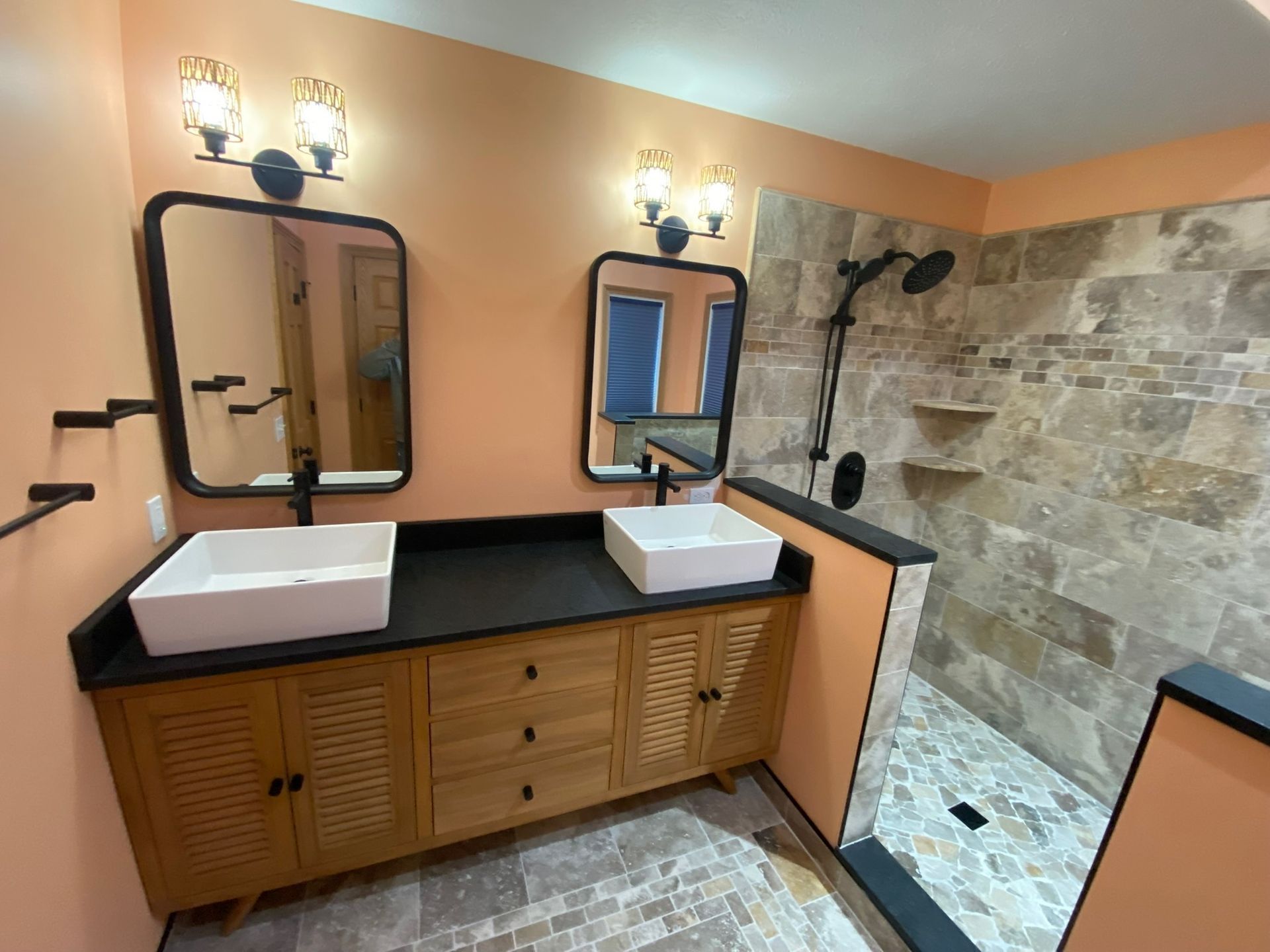 Bathroom vanity with two sinks, black countertop, wood cabinets, mirrors, and a stone-tiled shower wall