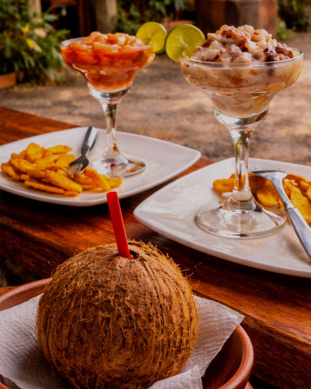 Dos cócteles de ceviche y una bebida de coco en una mesa de madera cerca de una playa, con chips de plátano.