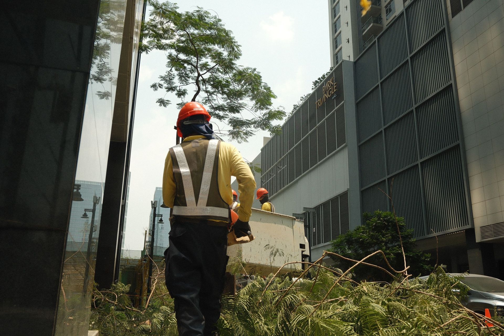 Worker in orange hard hat and reflective vest walking through overgrown area beside industrial buildings