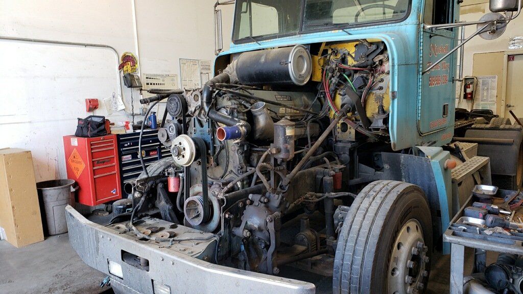 A semi-truck with its hood open, revealing the engine. It's in a repair shop, with tools and parts visible.