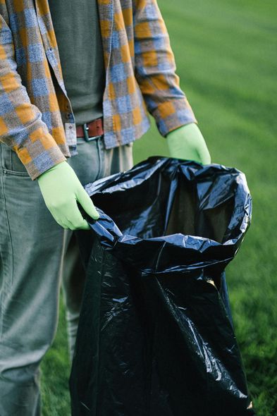 Person holding open a black trash bag while wearing green gloves outdoors.
