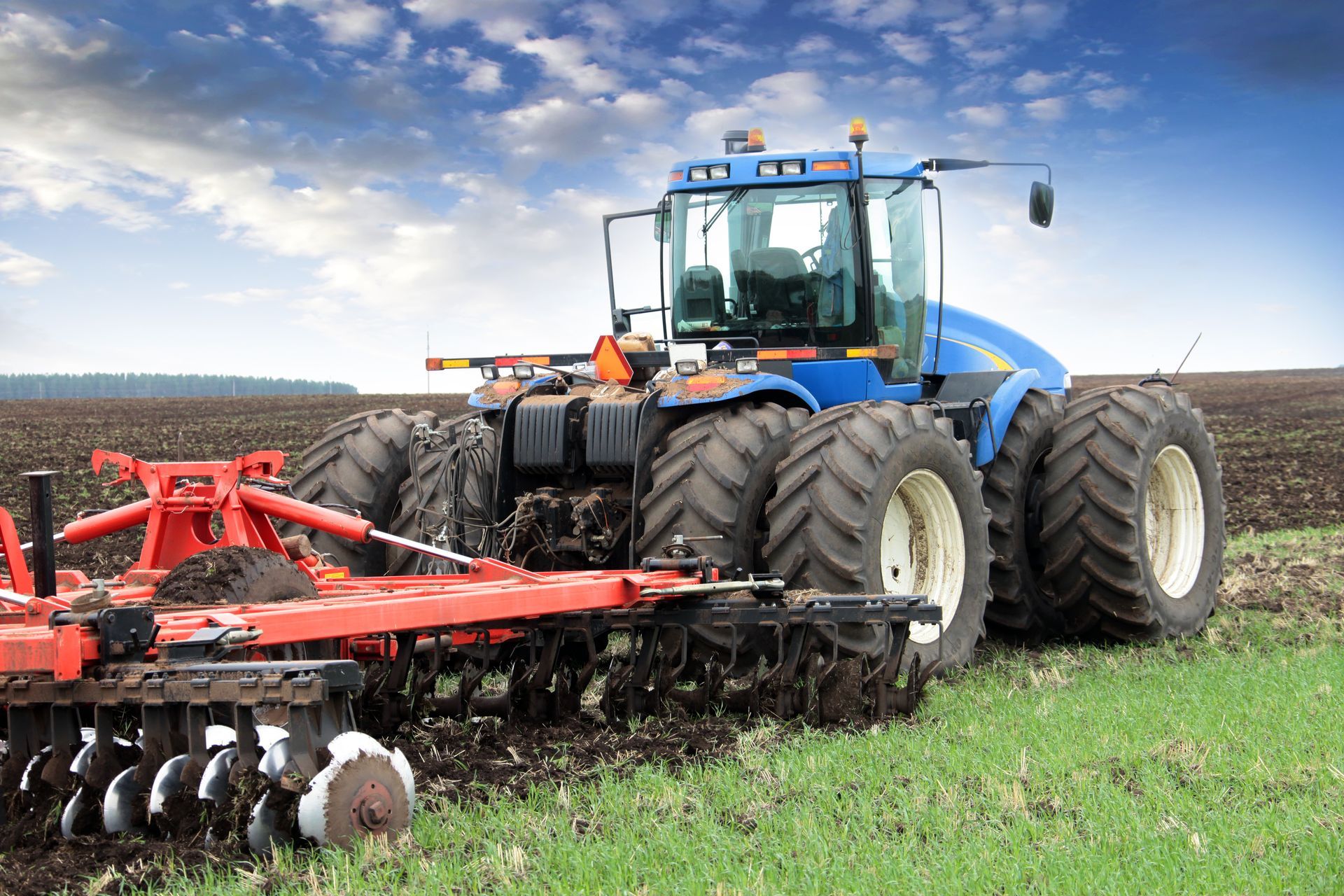 Blue tractor plowing a field with red tillage equipment under a cloudy sky
