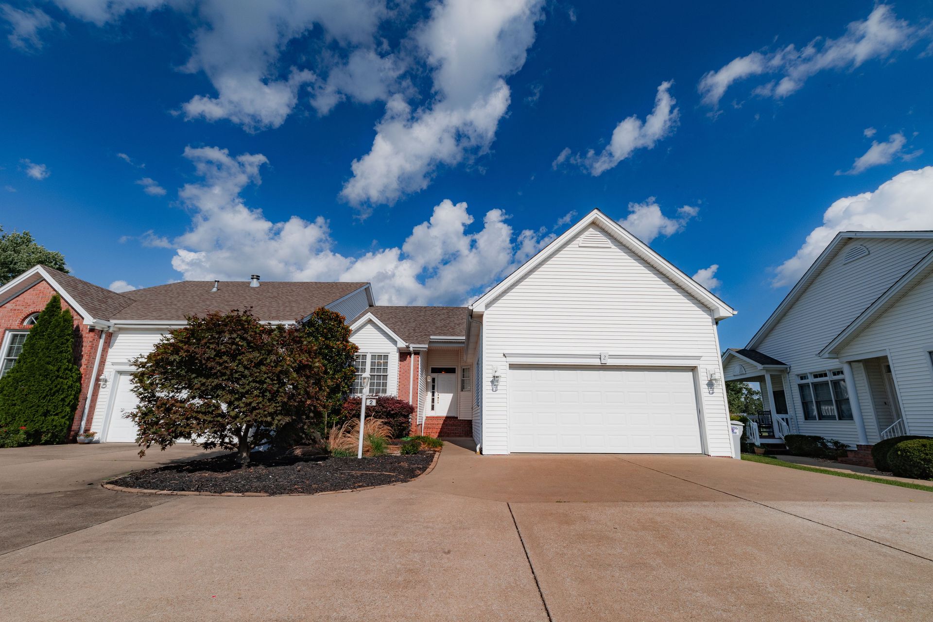 Suburban house with white garage, brick facade, and driveway under a blue sky with clouds