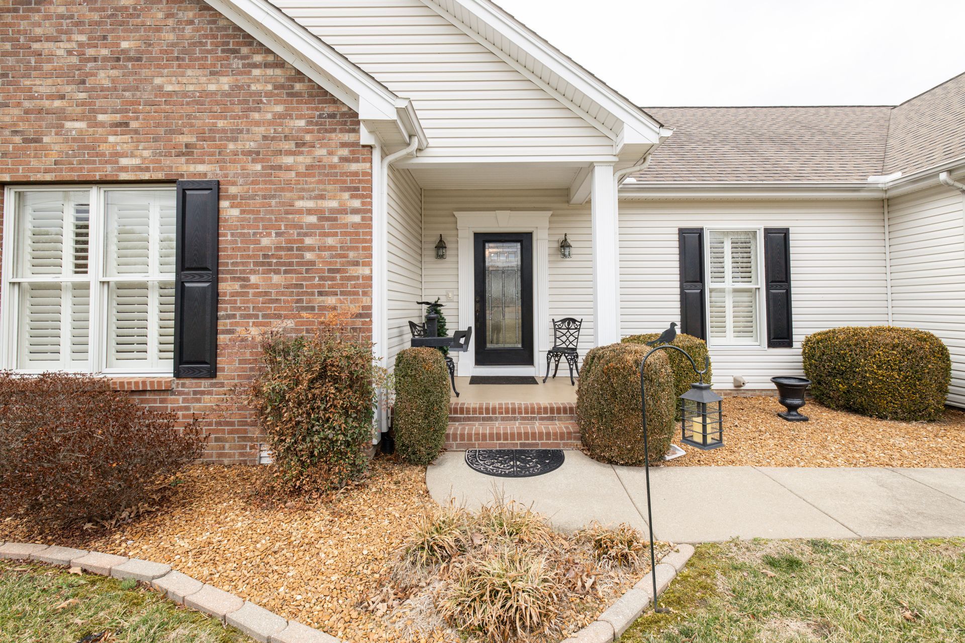 Front porch of a brick-and-siding house with white shutters and a concrete walkway.