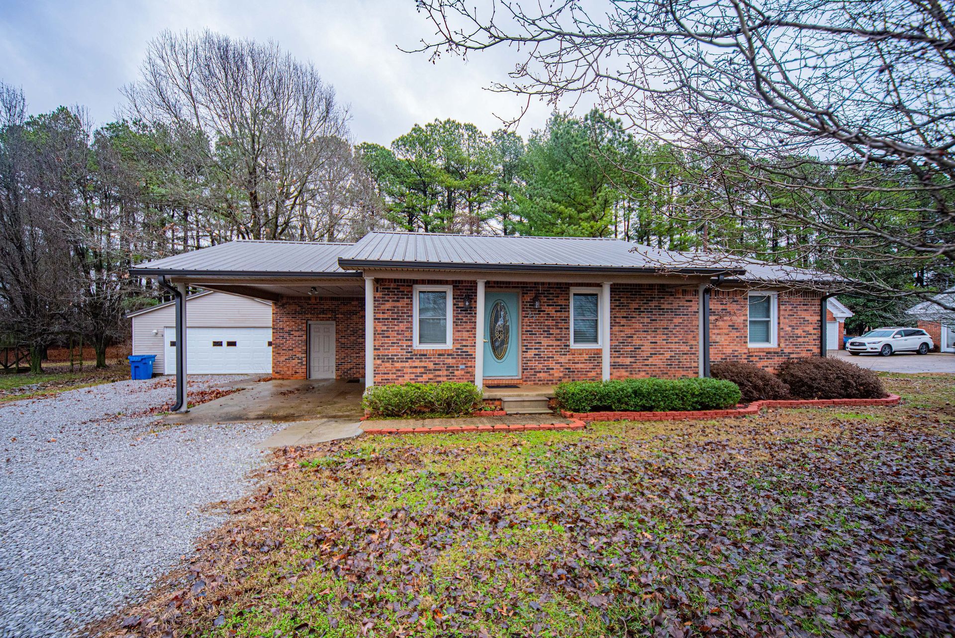 Brick ranch house with a white garage, front porch, and gravel driveway in a wooded yard