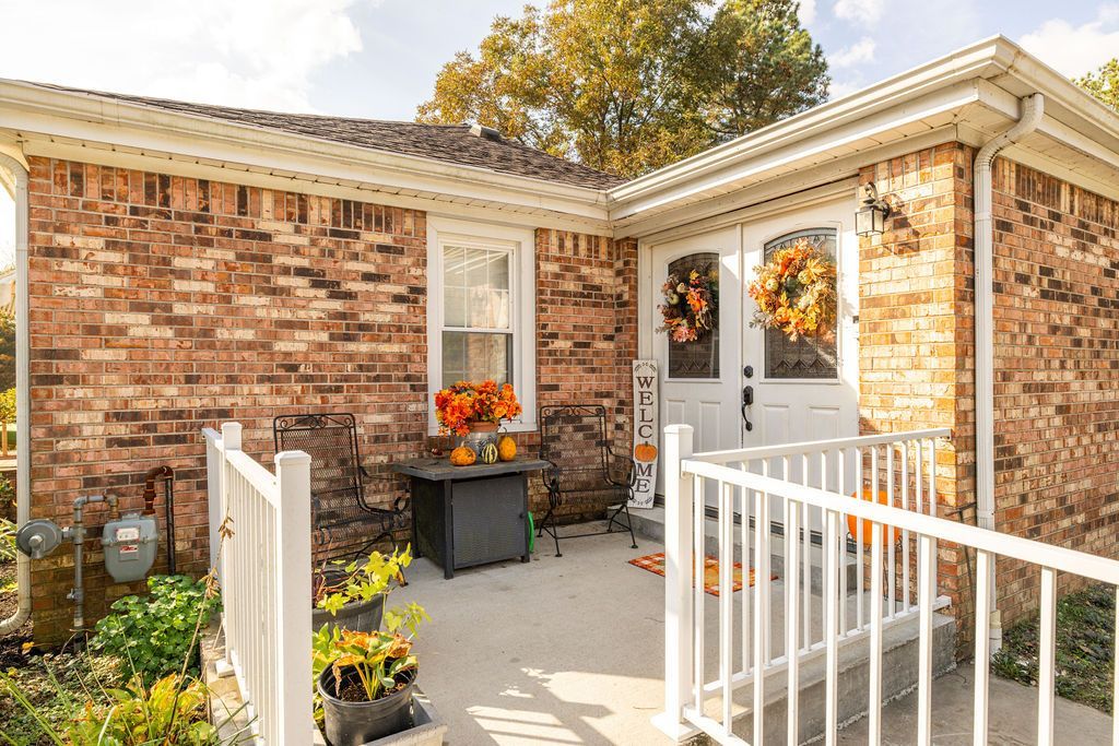 Brick house entrance with white railings, potted flowers, and autumn decorations on a sunny porch.