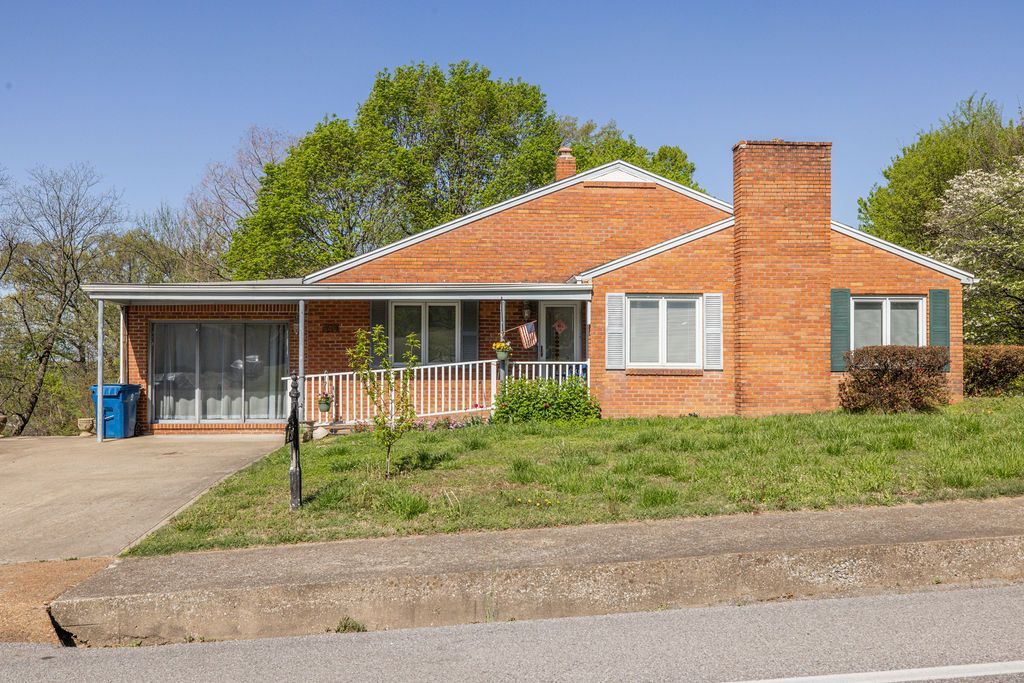 Single-story brick house with a carport, white trim, and a sloped gravel driveway on a sunny day