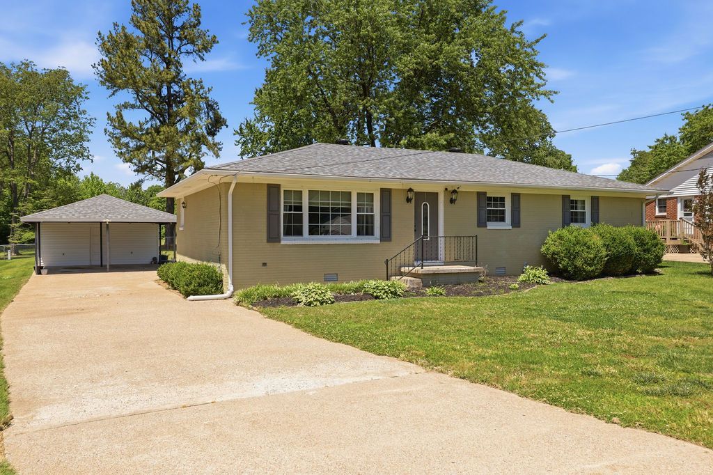 Single-story tan house with a long driveway, detached garage, and green lawn on a sunny day