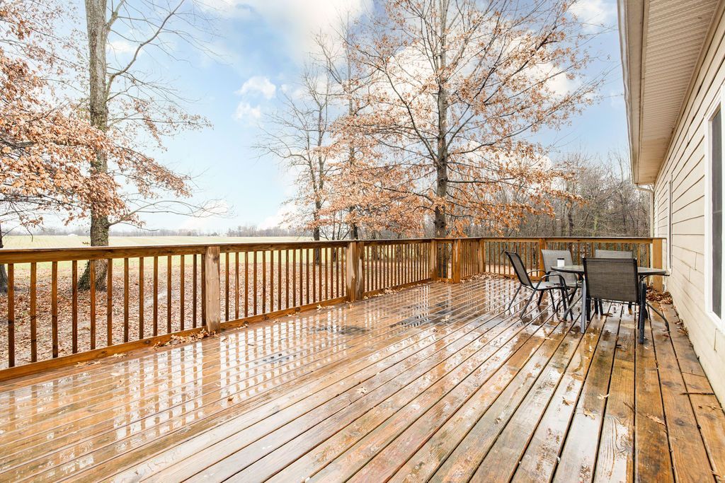 Wooden deck with railing and patio chairs, overlooking bare trees under a bright sky.