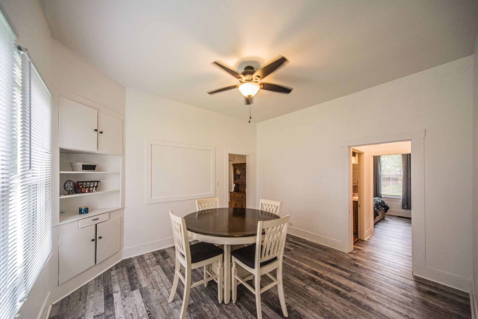 Bright dining room with a round table, white chairs, ceiling fan, and open doorway to another room.