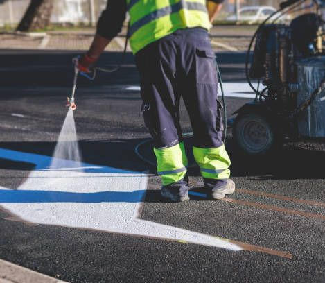 Road worker paints white lines on asphalt with a spray gun, wearing a reflective vest.