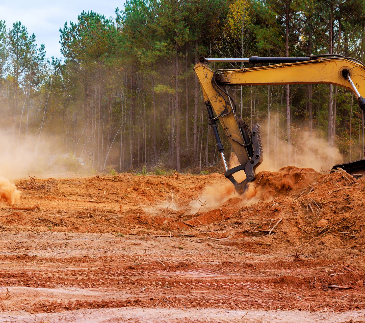 Yellow excavator clearing land, kicking up dust; forest in the background.