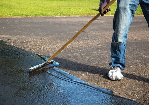 Person applying black sealant to a driveway with a squeegee tool.