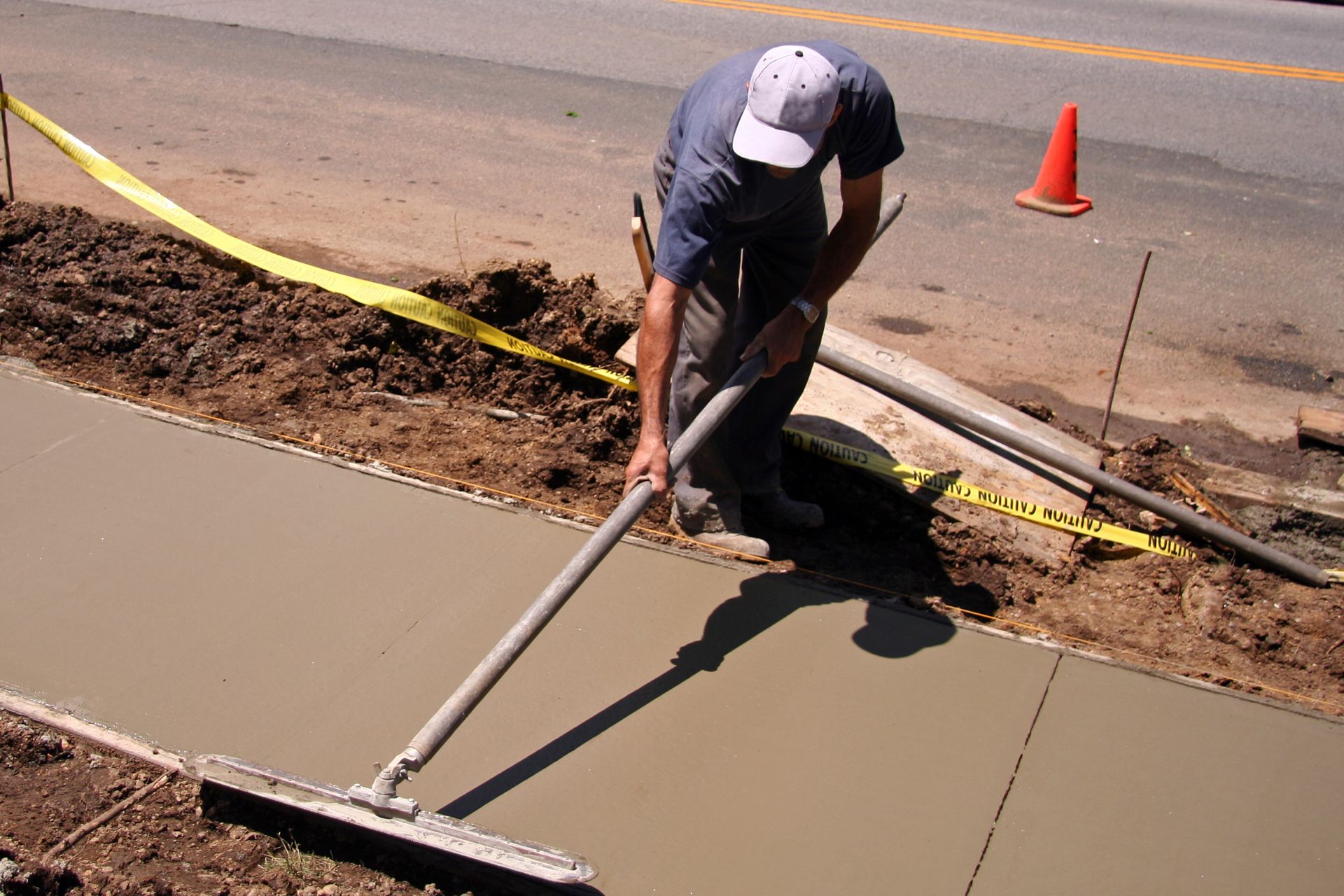 Construction worker smoothing wet concrete sidewalk with a squeegee tool. Yellow caution tape and orange cone visible.