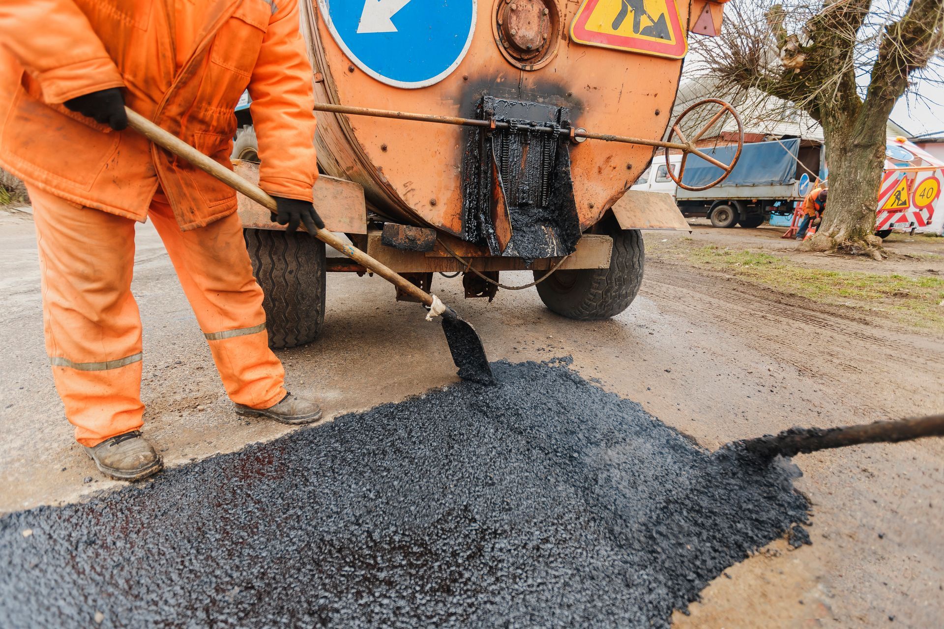 Road worker in orange suit spreading asphalt with a shovel near a tank on wheels.