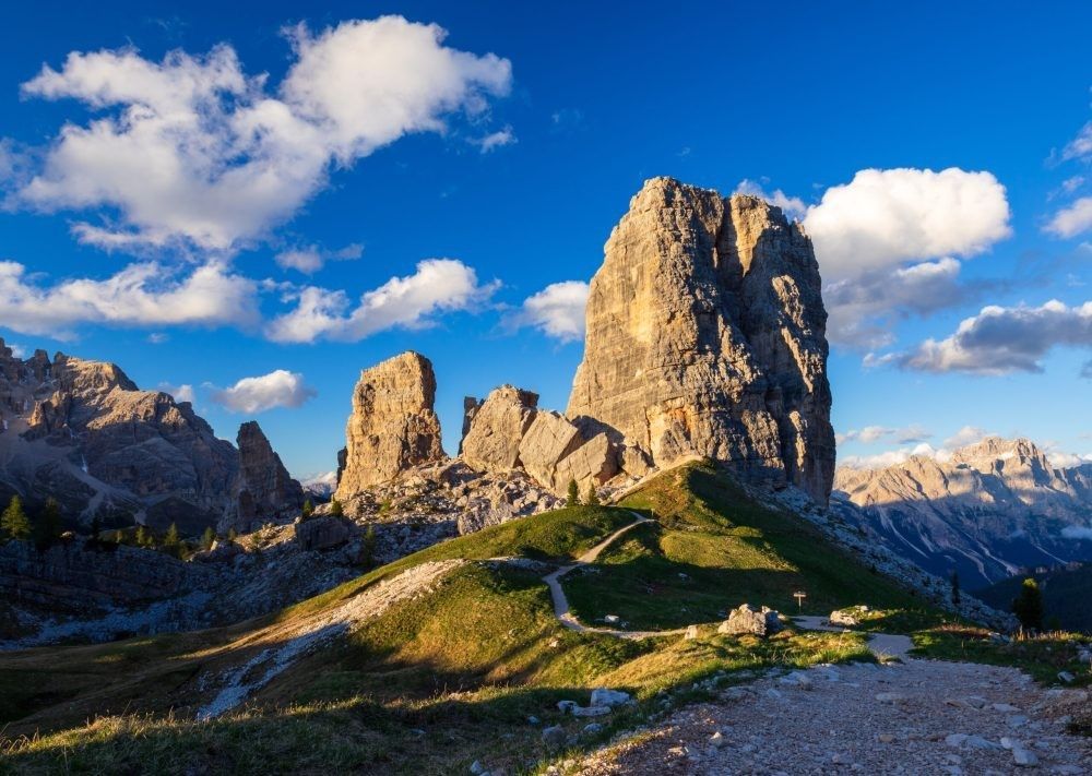 Jagged mountain peaks under a blue sky with clouds and a winding trail through green alpine terrain
