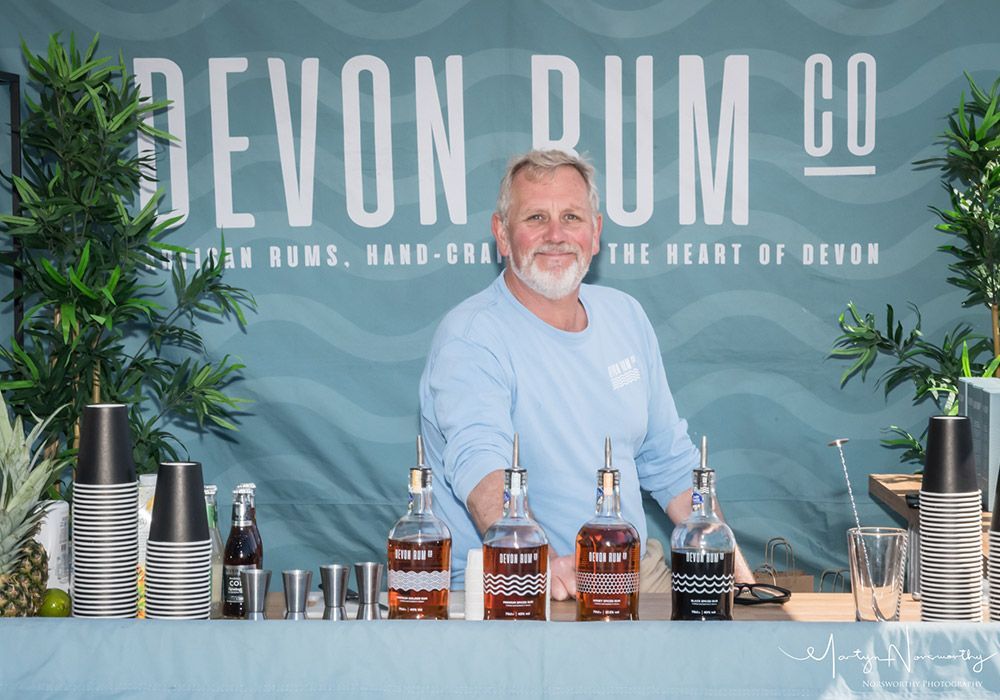 Man behind bar with Devon Rum Co. logo, bottles, and cocktail ingredients.