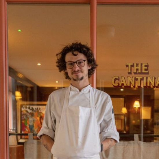 Chef stands in front of a restaurant, The Cantina. He wears a white apron and glasses.