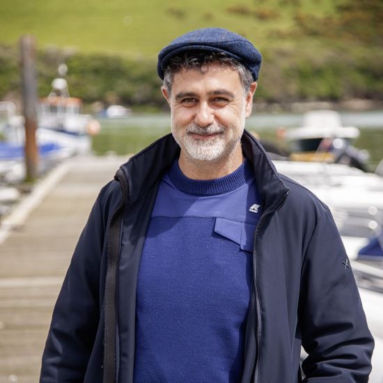 Man wearing a blue cap and sweater, standing on a pier near boats; smiling.