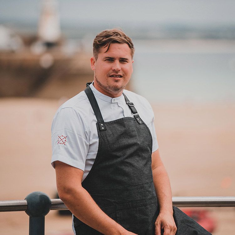 Chef in white shirt and black apron, posed outdoors near a beach, smiling.