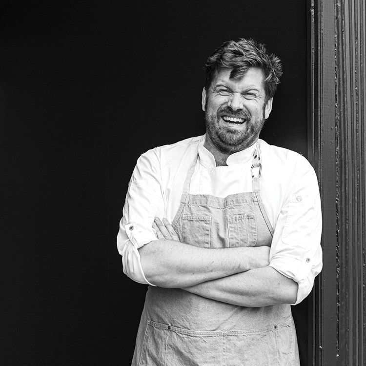 Man in chef's apron smiles, arms crossed, leaning against a dark doorway. Black and white.