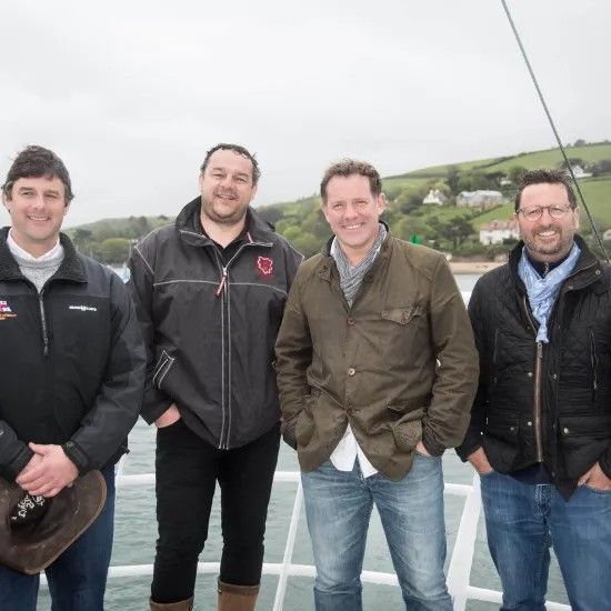 Four men on a boat, posing. Overcast day with water and green hills in the background.