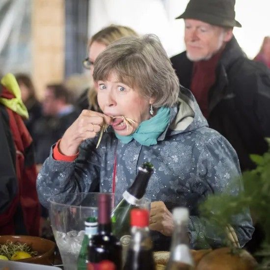 Woman with surprised expression eating food at an outdoor event, near bottles and people.