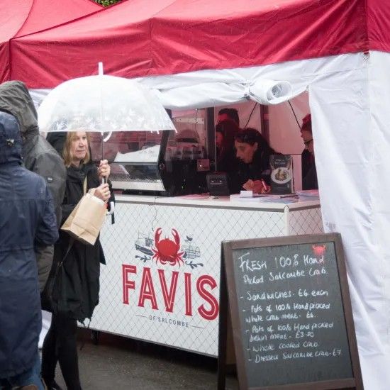 A woman with an umbrella shops at a crab food stall. The stall is red and white, with a chalkboard menu.
