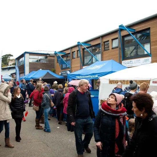People queueing at outdoor event with tents, building in background. Cloudy day.