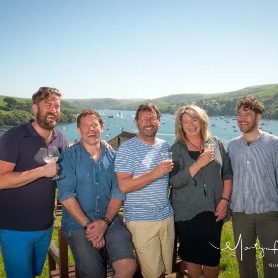 Five people smiling, holding glasses of wine, overlooking a scenic bay.