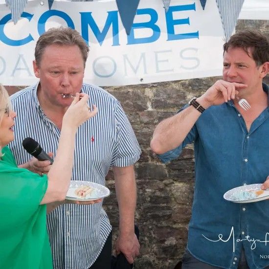 Woman feeding cake to two men outdoors. Men have plates and forks, are blowing out their cheeks.