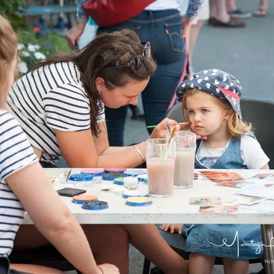 Woman paints face of a child at a table with paints and drinks.