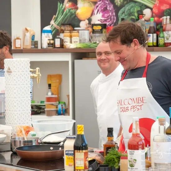 Chef at Salcombe Crab Fest cooking demonstration, wearing apron, smiling, with assistant.