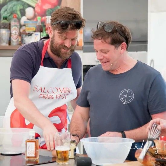 Two men smiling while cooking together, one wearing an apron that reads 