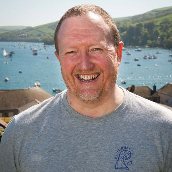 Man smiles broadly, outdoors near boats on a bay, wearing gray t-shirt.