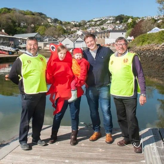 Group poses by water: two men in vests, a woman in a red octopus costume holding a child, and a man in jeans.
