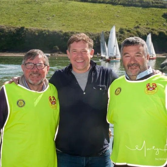 Three men in front of sailboats on water; two wearing yellow vests, smiling.